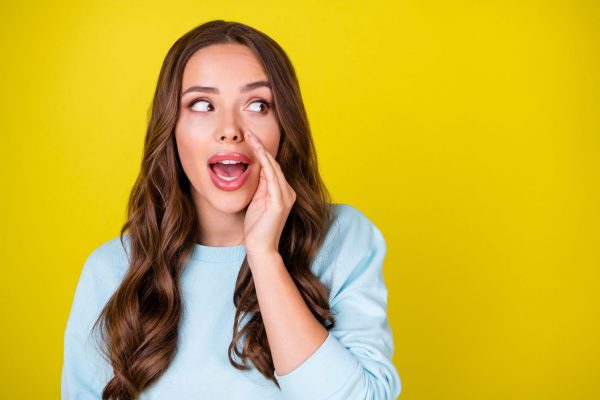 A white woman with brown hair whispering a secret, cupping her hand around her mouth, against a yellow background, symbolizing confidentiality and trust.